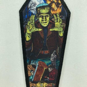 Könnte beinhalten: Ein Sarg-förmiger Aufnäher mit einem Frankenstein-Motiv. Das Monster hat grüne Haut, einen braunen Mantel und ausgestreckte Hände vor dem Hintergrund eines Vollmonds, Bäumen und Flammen. Der untere Teil des Aufnähers zeigt einen Sarg und die Worte "HUMAN PARTS".
