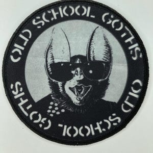 May include: A circular embroidered patch featuring a black and white graphic of a bat wearing sunglasses. The text "OLD SCHOOL GOTHS" is printed in a circular border around the central bat illustration. The patch has a black stitched border.