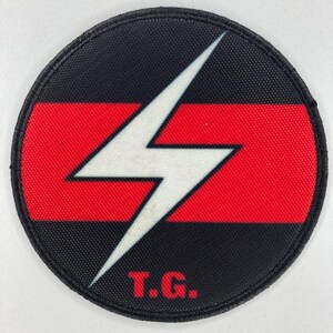 Puede incluir: Parche redondo con fondo negro y franja horizontal roja. Un rayo blanco está centrado. Las letras "T.G." están impresas en rojo debajo del rayo.