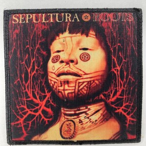 Puede incluir: Un parche cuadrado con el texto "SEPULTURA ROOTS" en rojo y naranja. El parche representa un retrato estilizado con marcas tribales y patrones rojos en forma de venas sobre un fondo oscuro. Tiene un borde negro.