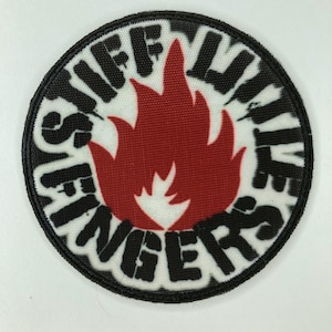 May include: A circular embroidered patch with a black border. The center features a red flame graphic on a white background. The black text "STIFF LITTLE FINGERS" is arranged in a circular pattern around the flame, following the edge of the patch.