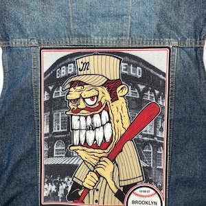 Puede incluir: Chaqueta vaquera azul con un parche grande que presenta un jugador de béisbol de dibujos animados sosteniendo un bate rojo. El parche incluye el texto "MELVINS" y "BROOKLYN N.Y." junto con la fecha "10-06-07".