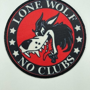 Peut inclure: Un écusson brodé circulaire avec un centre rouge et un graphique de tête de loup noir et blanc. La bordure noire contient le texte blanc LONE WOLF en haut et NO CLUBS en bas, séparés par quatre étoiles blanches de chaque côté.