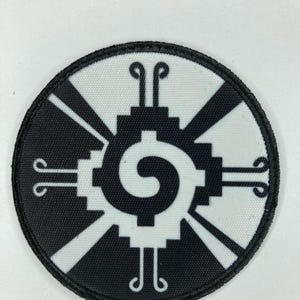 Puede incluir: Un parche bordado circular con un diseño geométrico en blanco y negro. El centro muestra un motivo de espiral rodeado por un patrón cuadrado escalonado, con cuatro brazos radiantes que terminan en pequeñas formas simétricas de voluta.