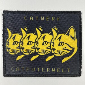 May include: A rectangular black patch with a stitched border featuring four yellow cat heads in a row. The text CATWERK is printed in yellow above the cats, and the text CATPUTERWELT is printed in yellow below the cats on the black fabric.