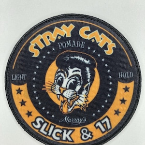 Pode incluir: Remendo circular com as palavras "Stray Cats Pomade" em letras amarelas sobre um fundo preto. No centro, um gato de desenho animado com cabelo penteado para trás. As palavras "Slick & 17" e "Murray's" também são visíveis.