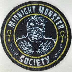 Könnte beinhalten: Ein runder, schwarzer Aufnäher mit gelbem Rand und Text. Der Aufnäher zeigt eine Mumienillustration in Weiß und Schwarz. Der Text lautet "MIDNIGHT MONSTER SOCIETY". Das Design enthält ein Ankh-Symbol.