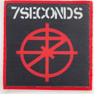 May include: A square fabric patch with a black background and a red border. The text 7SECONDS is printed in white at the top. Below the text is a red circular symbol featuring a cross and intersecting lines, resembling a stylized target design.