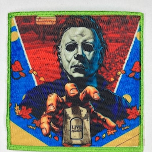 Puede incluir: Un parche con borde verde con una imagen colorida de una figura enmascarada sosteniendo una lápida que dice "RIP LIVE AGAIN". La figura lleva una chaqueta azul y negra y una máscara blanca con ojos negros.