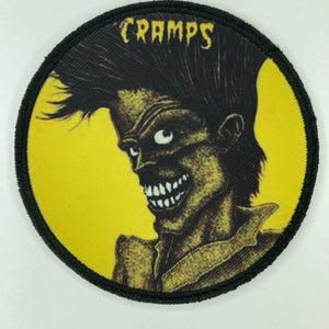 May include: A circular embroidered patch featuring a yellow background and a black graphic illustration of a stylized figure with a tall hairstyle and a wide, toothy grin. The word CRAMPS is printed in yellow at the top of the circular design.
