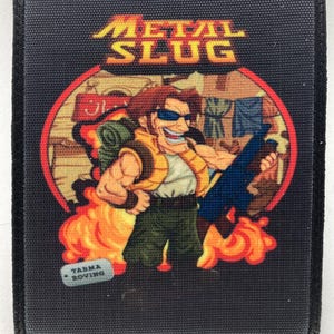 Op de afbeelding: Zwart rechthoekig embleem met het "METAL SLUG" logo in geel en oranje. Het embleem toont een stripfiguur met een zonnebril, die een wapen vasthoudt en omringd is door vlammen. De tekst "TARMA ROVING" is ook zichtbaar.