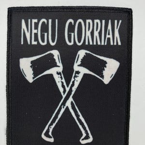 Puede incluir: Parche cuadrado negro con el texto blanco "NEGU GORRIAK" sobre una ilustración blanca de dos hachas cruzadas. Las hachas tienen hojas blancas y mangos gris claro. El parche tiene un borde negro.