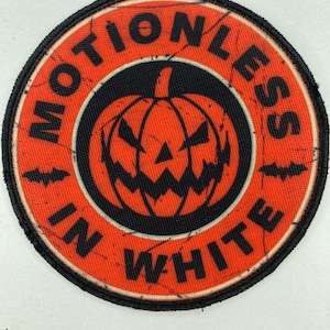 Puede incluir: Parche circular con fondo naranja y borde negro. Presenta una calabaza de Halloween en el centro y las palabras "MOTIONLESS IN WHITE" en una fuente desgastada. Dos siluetas de murciélagos negros en la parte superior.
