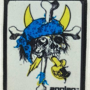 Puede incluir: Un parche rectangular con un diseño de calavera con un pañuelo azul, huesos cruzados y detalles amarillos. El diseño incluye un hacha amarilla y el texto "ZORLAC" en la parte inferior. El parche tiene un borde negro.