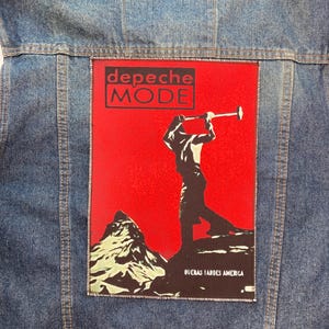 Pode incluir: Casaco de ganga azul com um patch retangular. O patch vermelho mostra o nome "Depeche Mode" numa caixa preta. Apresenta uma silhueta de uma pessoa com uma trombeta e o texto "Buenas Tardes America".