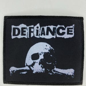 Puede incluir: Parche bordado en blanco y negro con la palabra "DEFIANCE" en una fuente desgastada y un diseño de calavera y huesos cruzados.