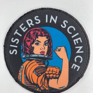 Puede incluir: Un parche redondo con las palabras "SISTERS IN SCIENCE" que rodean una ilustración retro de una mujer con un traje espacial naranja, flexionando su brazo. La mujer tiene el pelo rojo y está sobre un fondo azul.