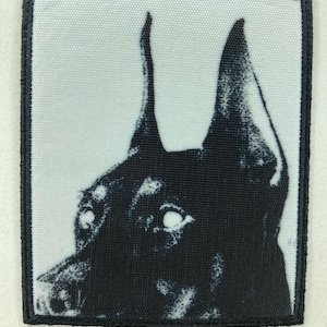 Puede incluir: Parche cuadrado en blanco y negro con borde negro que muestra la cabeza de un Doberman Pinscher. Las orejas del perro están erguidas y sus ojos son visibles.