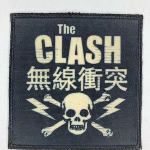 Puede incluir: Un parche cuadrado con fondo azul oscuro presenta el texto "The CLASH" en crema, junto con caracteres japoneses. Debajo, una calavera y huesos cruzados de color crema se cruzan con rayos.