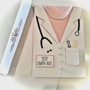 May include: A greeting card shaped like a doctor's coat, with a stethoscope, syringe, and name tag that reads "TEST SMITH, M.D." The coat is white with pink accents and a pearl necklace. A white box with a decorative bow is on the left.