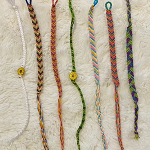Peut inclure: Sept bracelets d'amitié colorés avec un motif en chevron et un charme en forme de tournesol. Les bracelets sont faits de fil à broder et sont noués à l'extrémité.