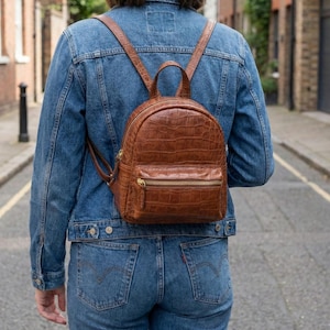 Puede incluir: Una mochila de cuero marrón con textura de cocodrilo, con asa superior y correas ajustables. La mochila tiene un bolsillo frontal con cremallera y herrajes dorados. La persona lleva una chaqueta vaquera azul y vaqueros.