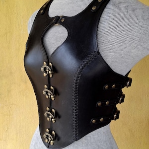 May include: A black leather corset with a heart-shaped cutout in the front. The corset has a series of decorative buckles and straps along the sides and front.