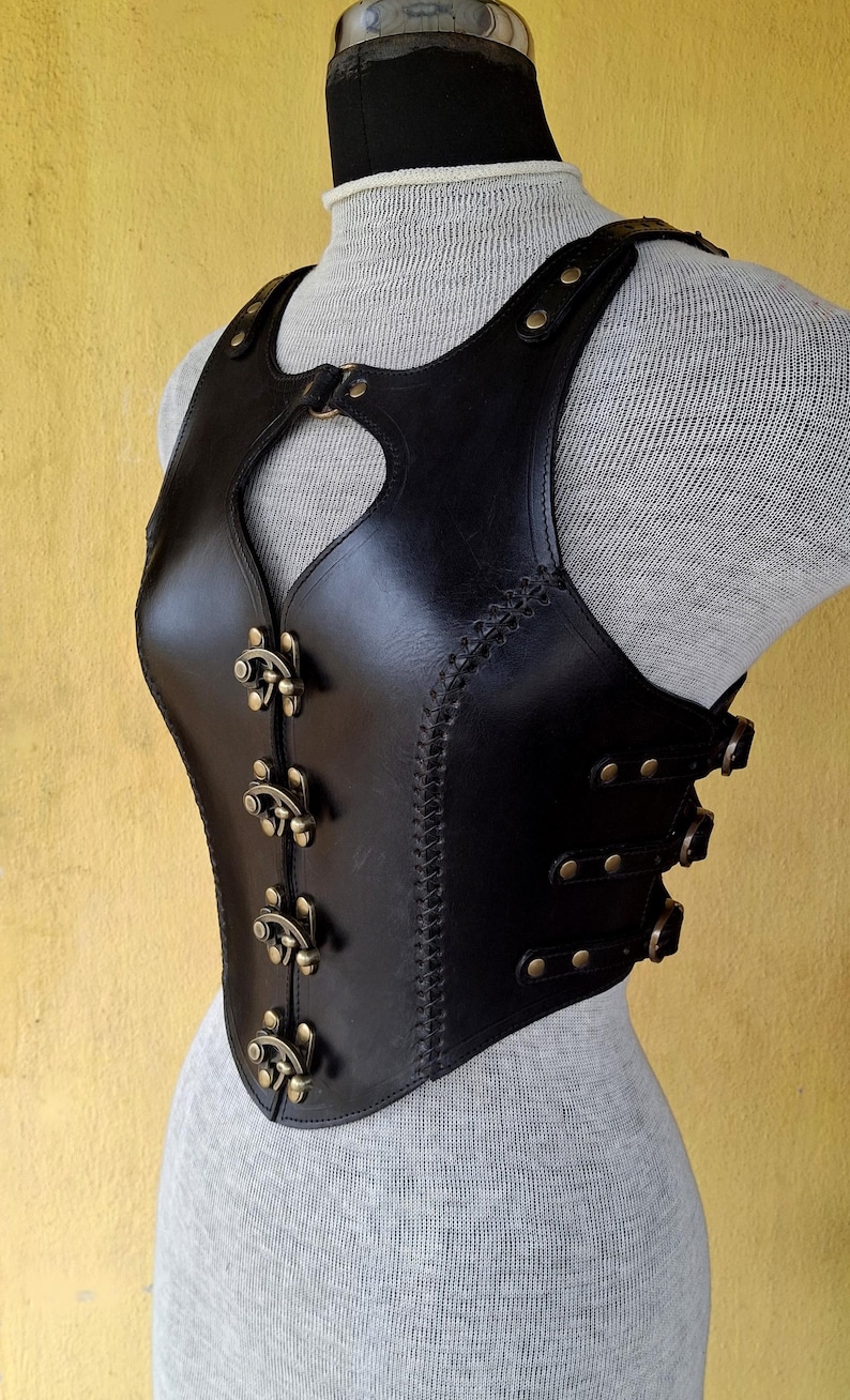 May include: A black leather corset with a heart-shaped cutout in the front. The corset has a series of decorative buckles and straps along the sides and front.
