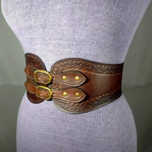 May include: A brown leather belt with a decorative embossed design and double buckles. The belt has a wide, curved shape and is secured with brass-toned hardware.