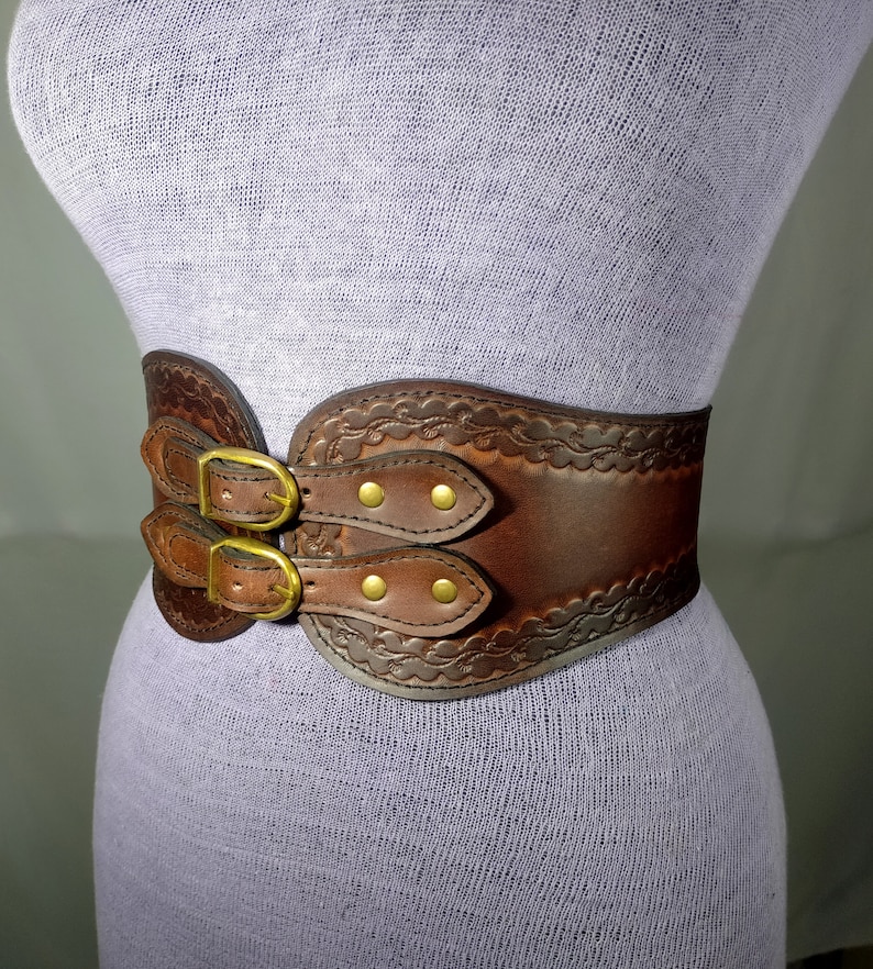 May include: A brown leather belt with a decorative embossed design and double buckles. The belt has a wide, curved shape and is secured with brass-toned hardware.