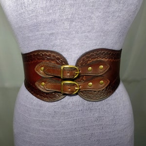May include: A brown leather belt with two double buckles and decorative stitching. The belt has a wide, curved shape and is fastened with brass hardware.