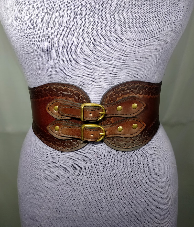 May include: A brown leather belt with two double buckles and decorative stitching. The belt has a wide, curved shape and is fastened with brass hardware.