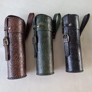 May include: Three leather quivers in brown, green, and black, each with a floral embossed pattern. They have a strap and buckle closure, and a handle. The quivers are cylindrical and appear to be handmade.