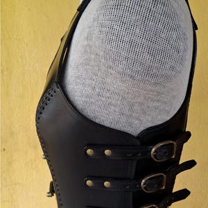 May include: Black leather vest with multiple buckles and straps. The vest has a decorative stitching detail and is worn on a mannequin.