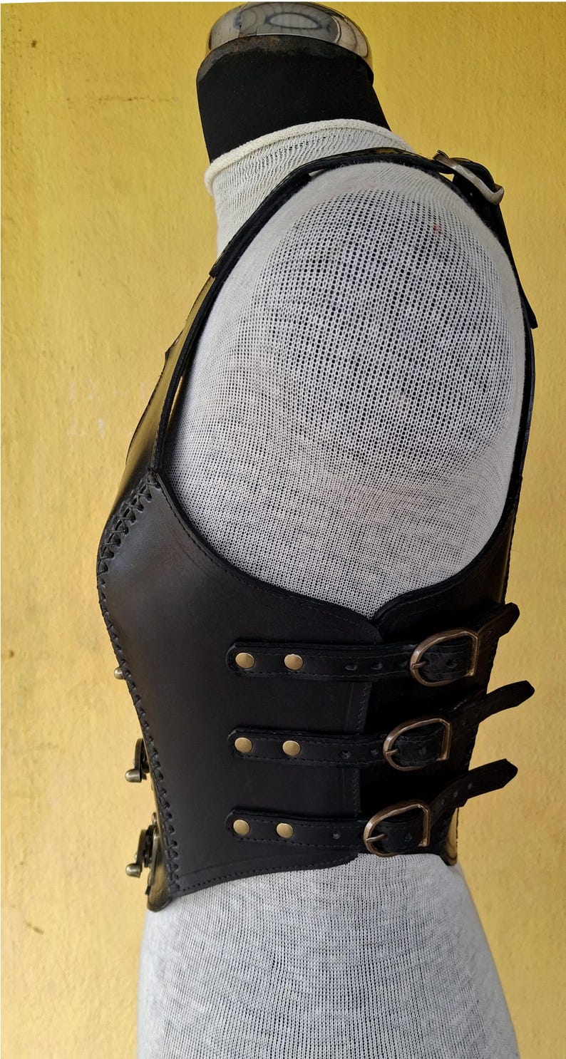May include: Black leather vest with multiple buckles and straps. The vest has a decorative stitching detail and is worn on a mannequin.