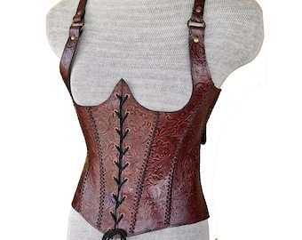 May include: A brown leather corset with a floral embossed pattern. The corset has adjustable straps and a lace-up front. The garment is displayed on a mannequin. The corset is approximately 30 cm in height.