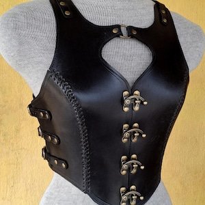 May include: A black leather corset with a heart-shaped cutout in the front. The corset has a series of brass buckles down the front and is stitched with black thread.