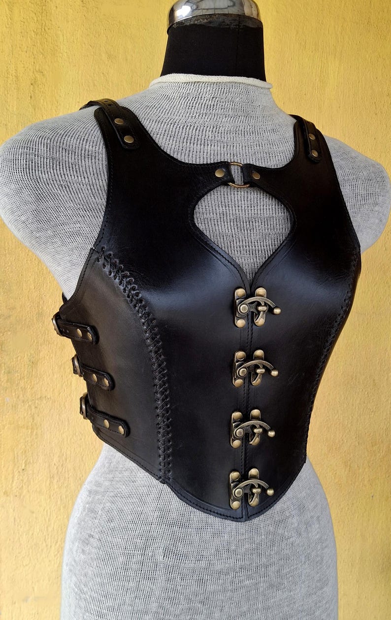 May include: A black leather corset with a heart-shaped cutout in the front. The corset has a series of brass buckles down the front and is stitched with black thread.