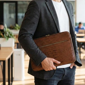 May include: A brown leather laptop case with a crocodile skin effect. The case has a gold zip and is held by a person wearing a grey blazer, white t-shirt, and blue jeans. The background shows an office setting.