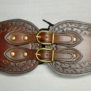 May include: A brown leather belt with two brass buckles and decorative stitching. The belt is designed to be worn around the waist.