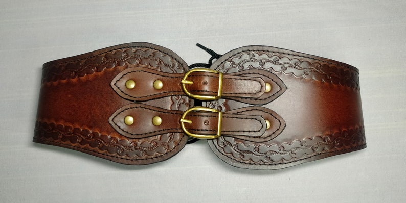 May include: A brown leather belt with two brass buckles and decorative stitching. The belt is designed to be worn around the waist.