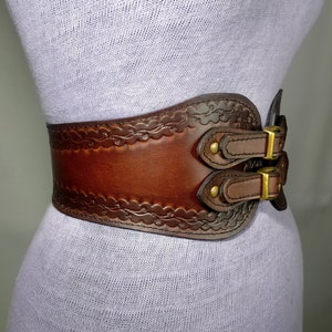 May include: A brown leather belt with a decorative embossed design and two double buckles. The belt is worn on a mannequin.