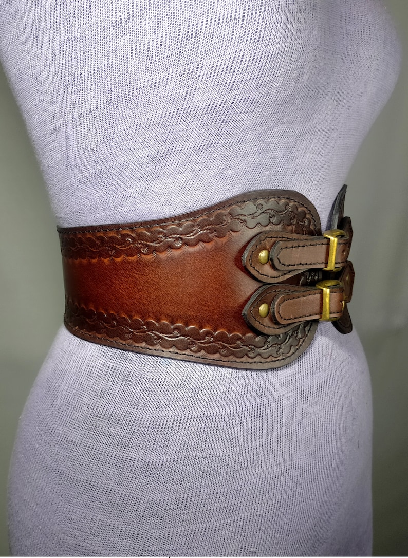 May include: A brown leather belt with a decorative embossed design and two double buckles. The belt is worn on a mannequin.
