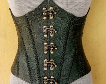 May include: A dark green leather corset with a floral embossed pattern. The corset features a pointed top and bottom, with decorative metal clasps down the front. The corset is designed to cinch the waist and has a vintage aesthetic.