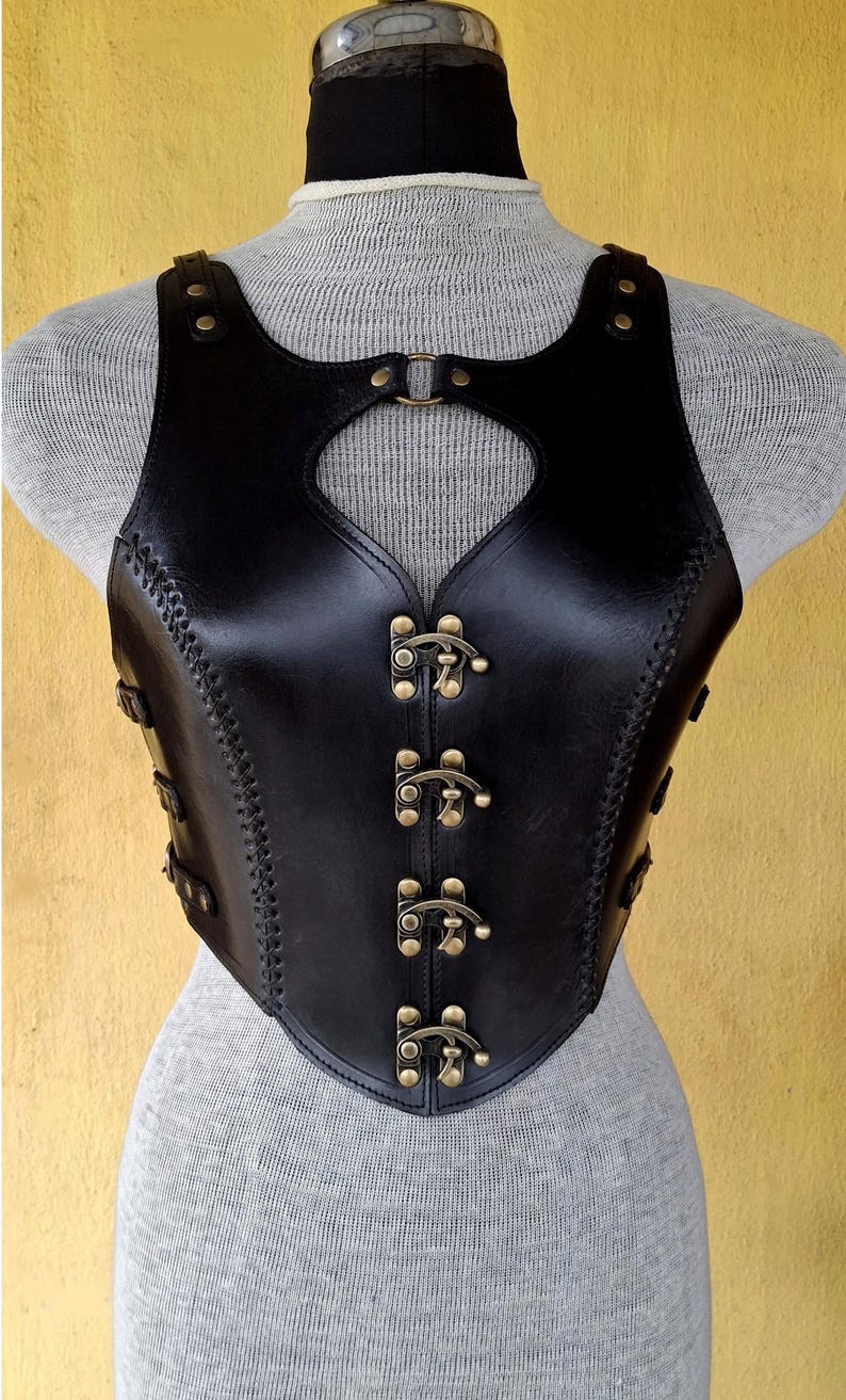 May include: A black leather corset with a heart-shaped cutout in the front. The corset has a series of decorative brass buckles down the front.