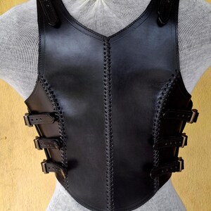 May include: Black leather vest with a decorative stitched design and multiple buckles. The vest has a V-shaped neckline and a fitted silhouette.
