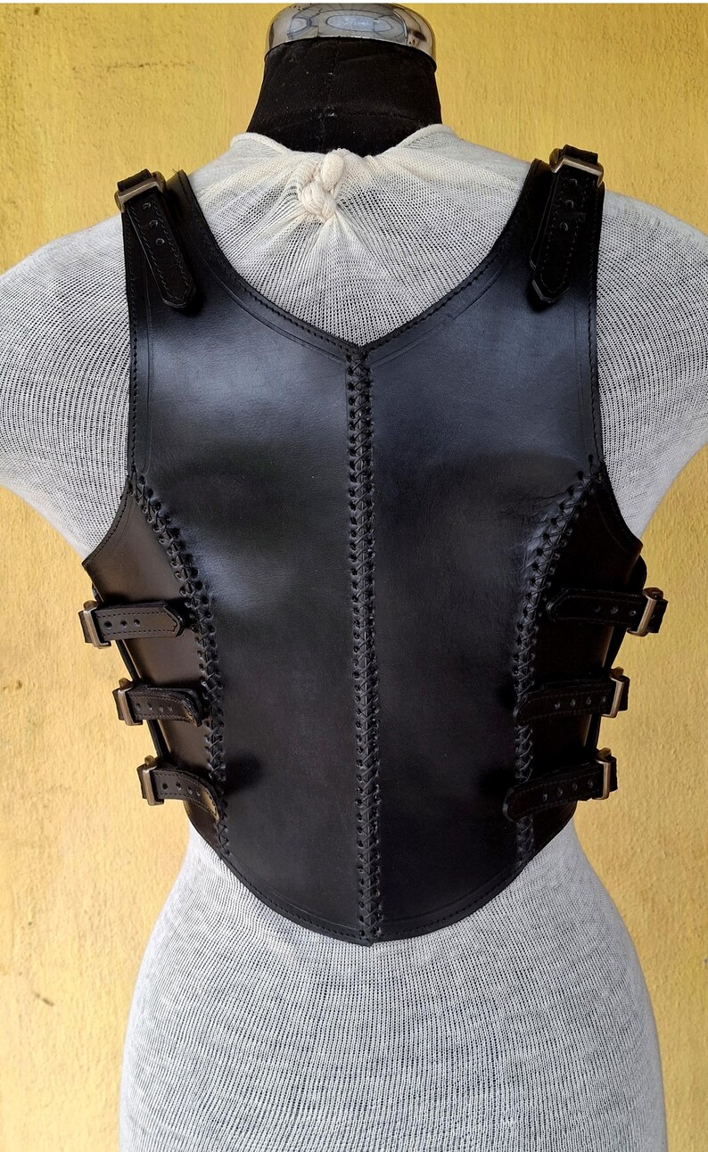 May include: Black leather vest with a decorative stitched design and multiple buckles. The vest has a V-shaped neckline and a fitted silhouette.