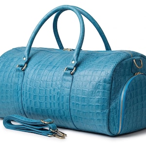 May include: A turquoise blue duffel bag with a crocodile skin texture. The bag has two top handles, a detachable shoulder strap, and a side pocket. The bag is made of leather and has gold-coloured zips and hardware.