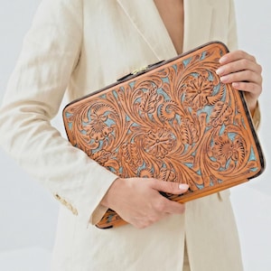 Könnte beinhalten: Eine rechteckige, braune Ledertasche mit floralem Prägemuster und türkisfarbenem Hintergrund. Die Tasche hat einen schwarzen Reißverschluss und wird von einer Person in einem cremefarbenen Blazer gehalten. Die Tasche ist wahrscheinlich für einen Laptop oder ein Tablet gedacht.