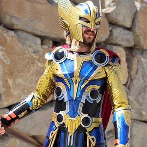 Può includere: Un costume cosplay dettagliato di un supereroe, con un elmo dorato con accenti blu, un'armatura blu e oro e un mantello rosso. Il costume presenta dettagli intricati e un aspetto metallico, suggerendo un design di alta qualità.